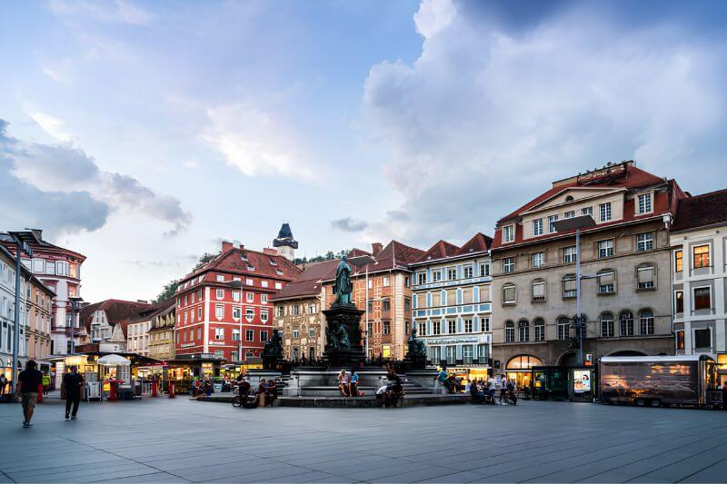 Hauptplatz rộng rãi với các dãy nhà cổ và Cột Chúa Ba Ngôi nổi bật ở trung tâm.